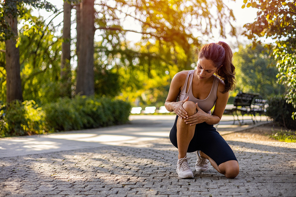 Young woman feeling pain in her knee. Woman runner got sports injury running on forest trail.